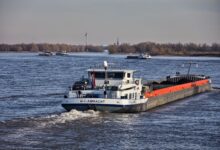 A large barge traveling along an inland waterway near an industrial terminal